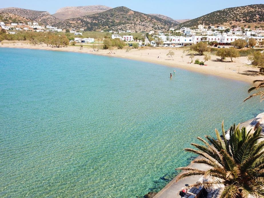 Scenic view of a beach with turquoise water, a sandy shore, and white buildings nestled against green hills. Palm trees frame the foreground.