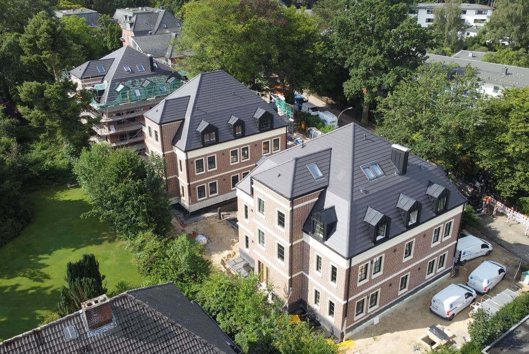 Aerial view of brick residential buildings with dark pitched roofs, scaffolding and vans, surrounded by dense trees and a green lawn