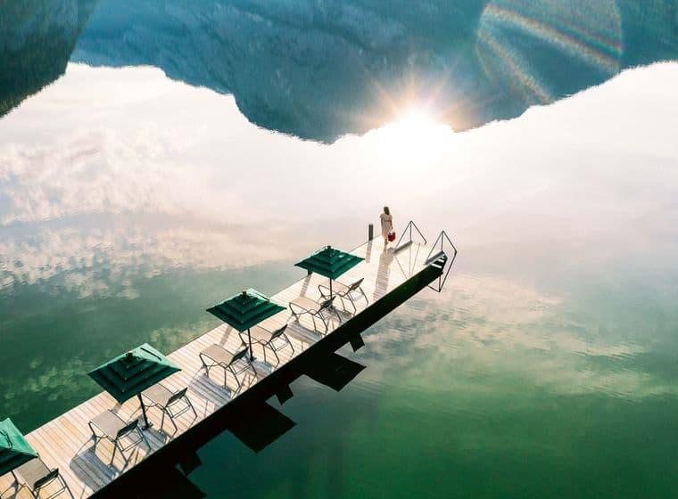 Vista aérea de un muelle de madera con sombrillas y sillas verdes en un lago, con una mujer de pie al final, montañas al fondo y el sol reflejándose en el agua.