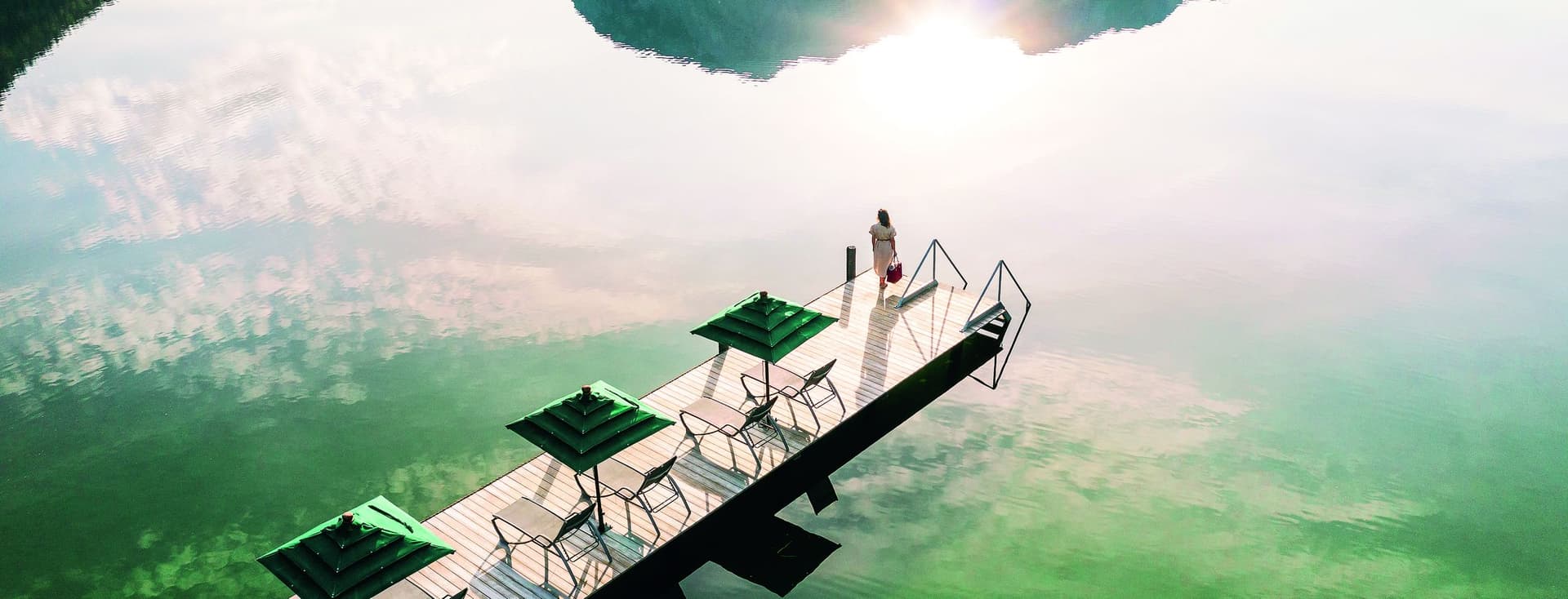 Une femme se tient sur un quai avec des parasols et des chaises verts, surplombant un lac qui reflète les montagnes et le soleil.