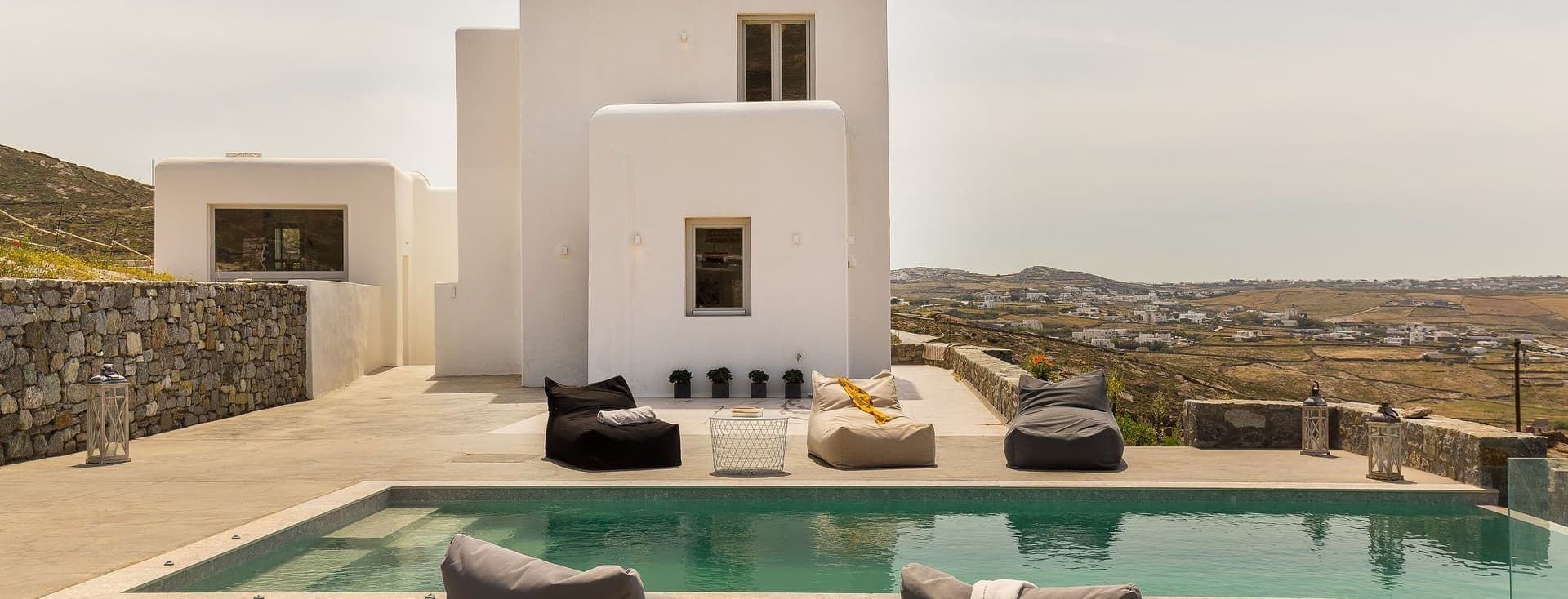 Luxury villa with a pool and bean bag chairs. White building with windows, and a view of the landscape.