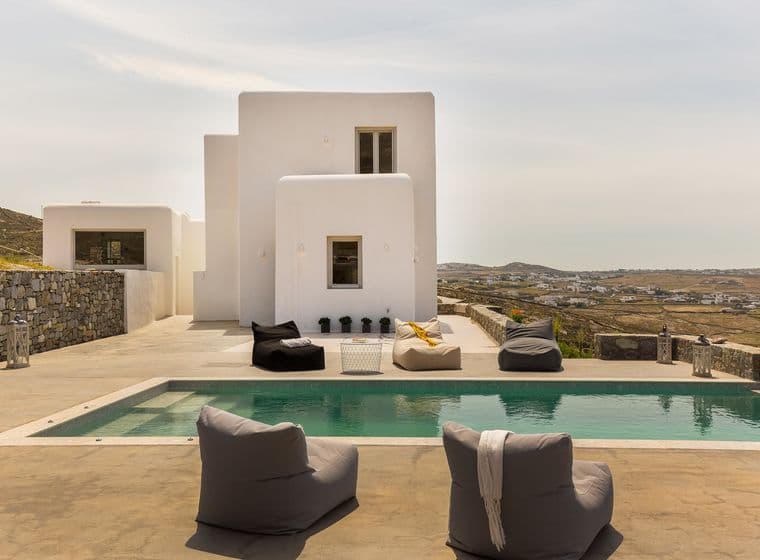Luxury villa with a pool and bean bag chairs. White building with windows, and a view of the landscape.