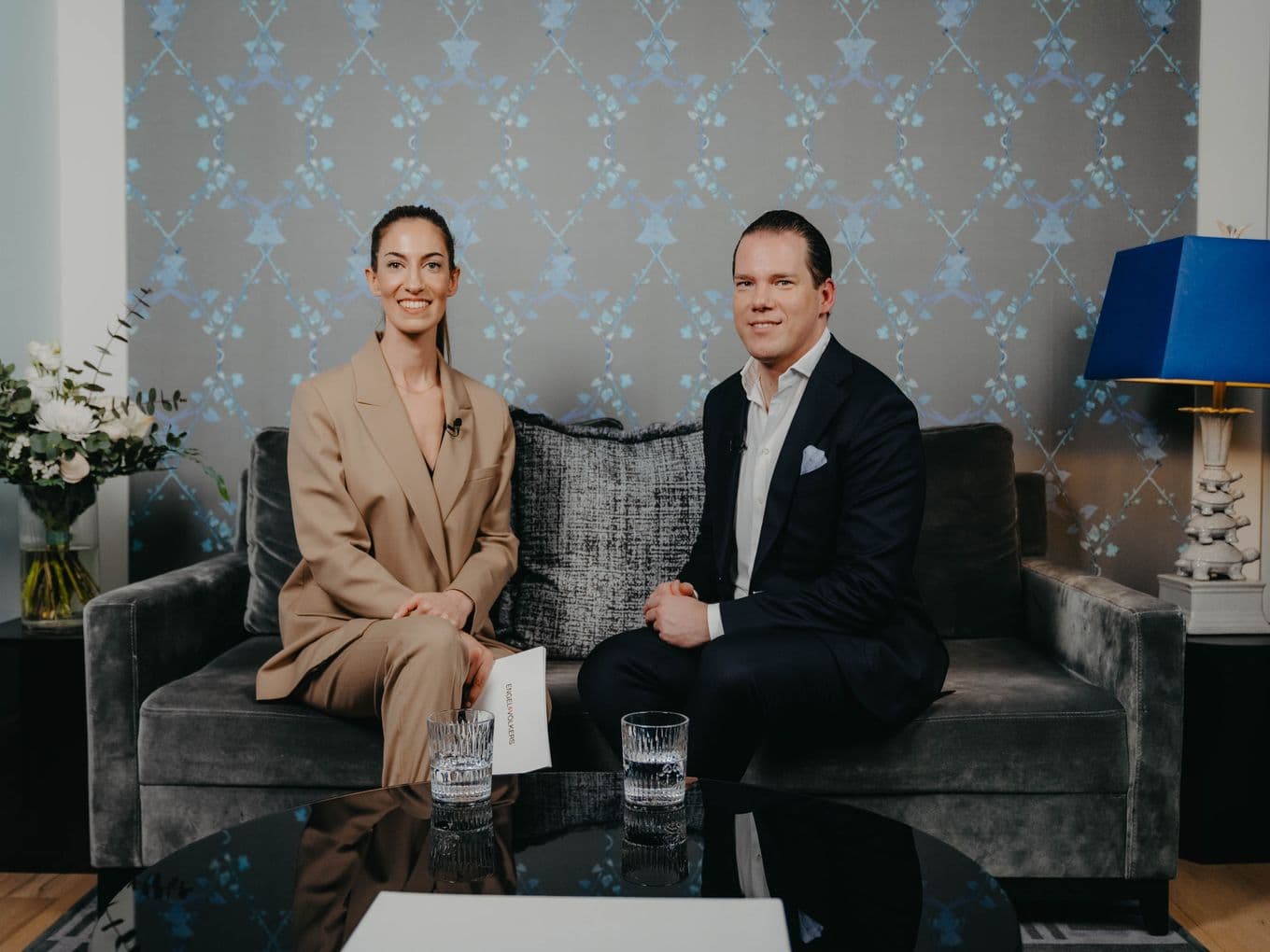Two people sitting on a sofa in a modern room with patterned wallpaper, a blue lamp, and a table with glasses of water.