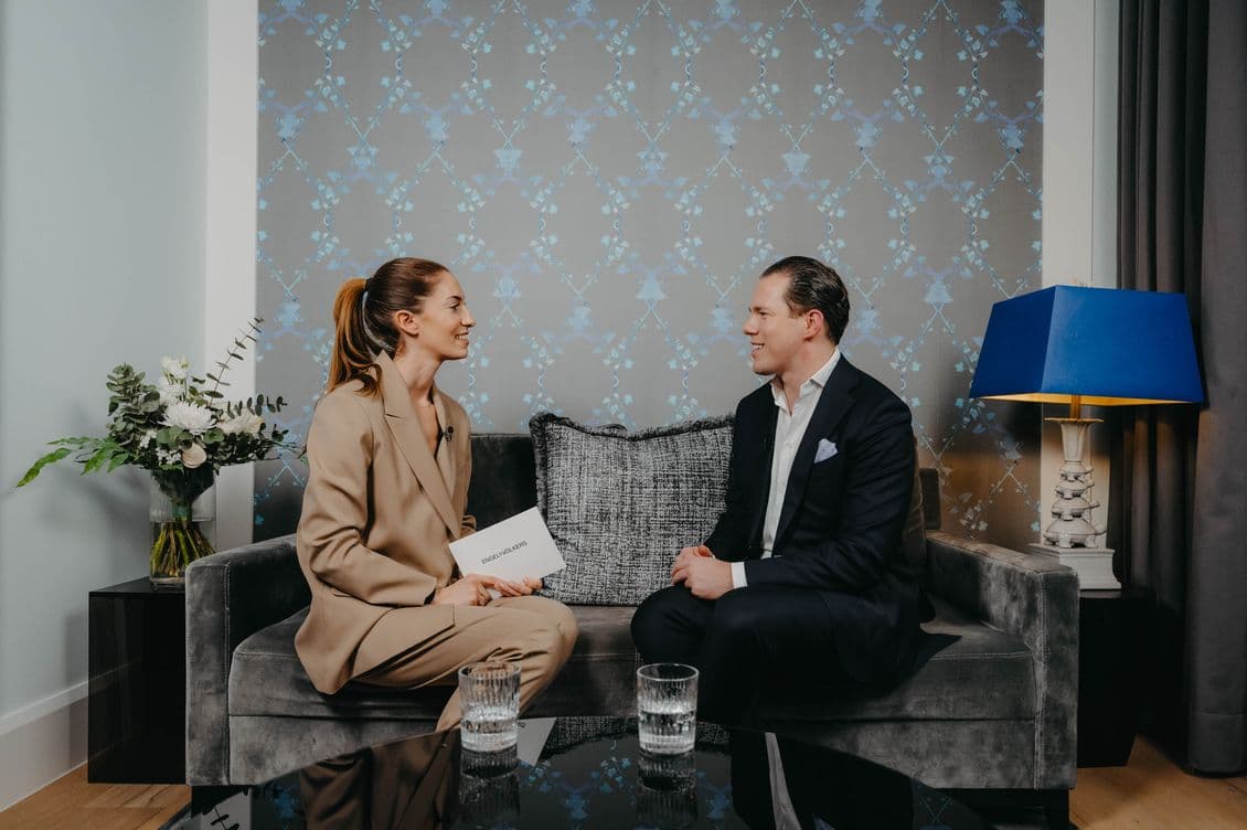 Two people sitting on a gray sofa in a modern room, engaged in conversation. A table with glasses of water is in front of them.