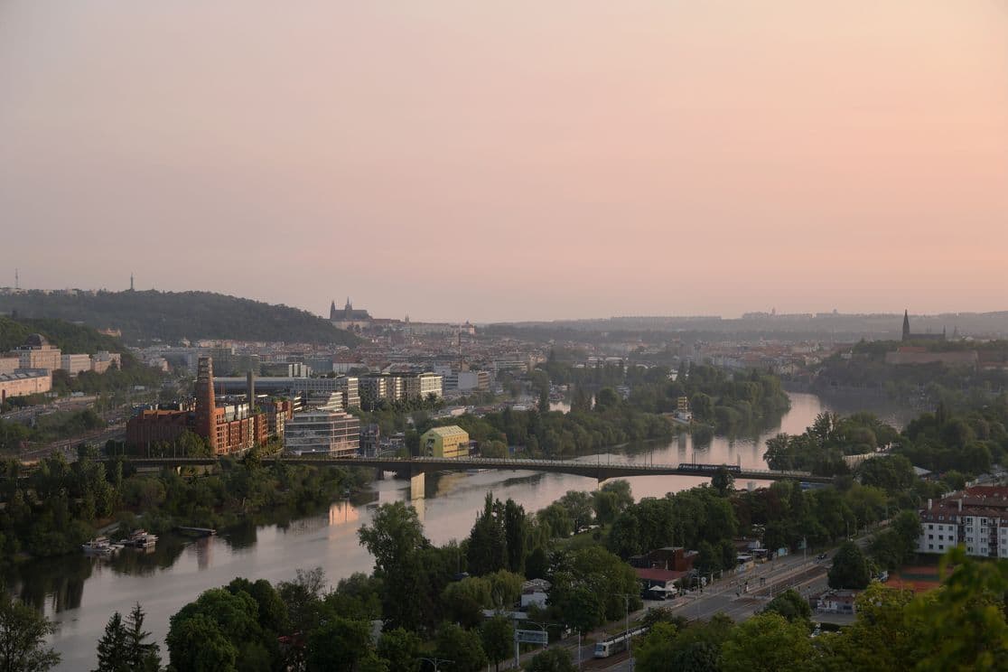 Panoramatický pohled na Prahu při západu slunce, v popředí nová rezidenční zástavba u řeky, v dálce Petřín, Pražský hrad a městská zástavba.