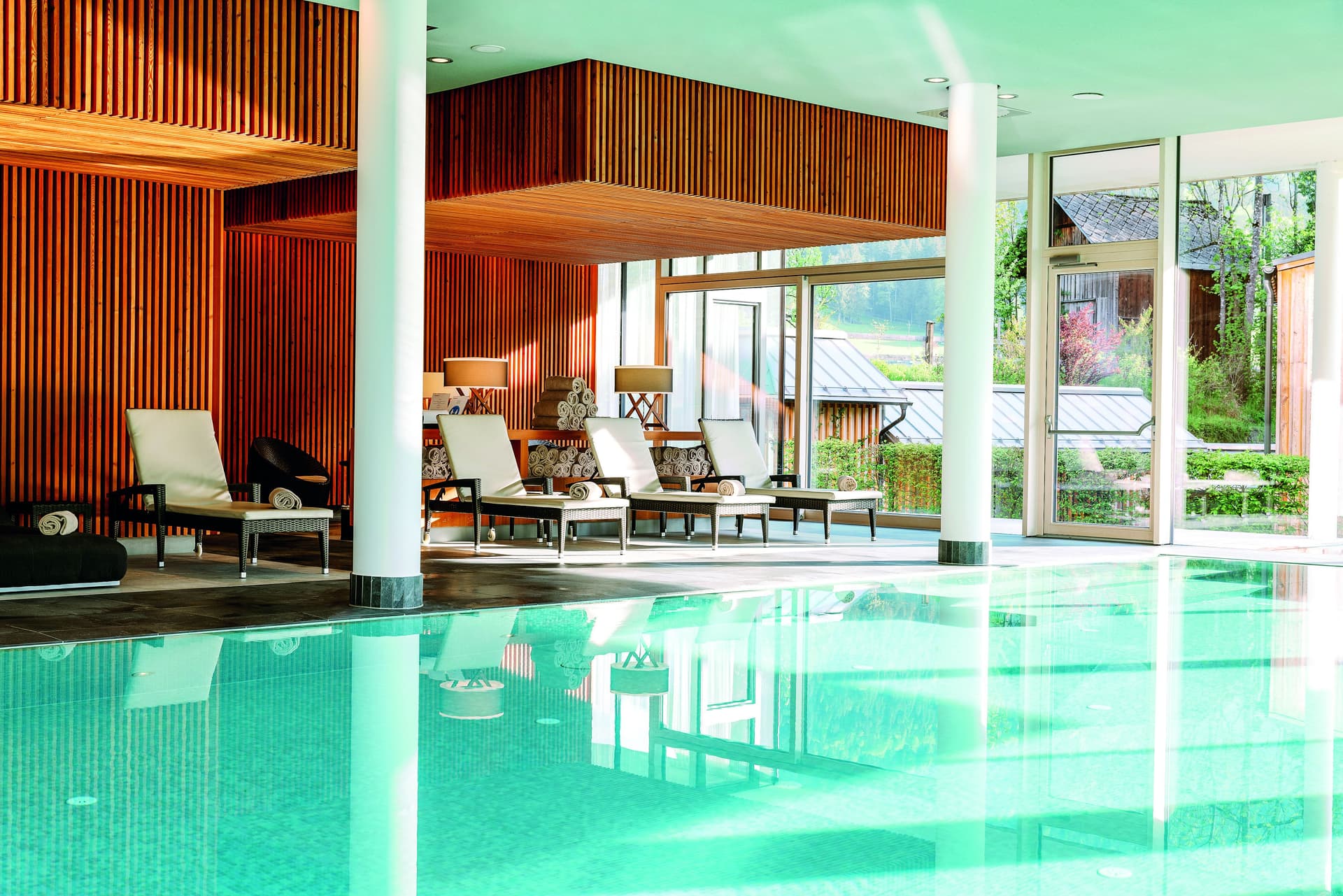 Espace piscine intérieure avec chaises longues, serviettes et murs en bois. De grandes fenêtres offrent une vue sur l'extérieur. L'eau de la piscine est d'une couleur turquoise claire.