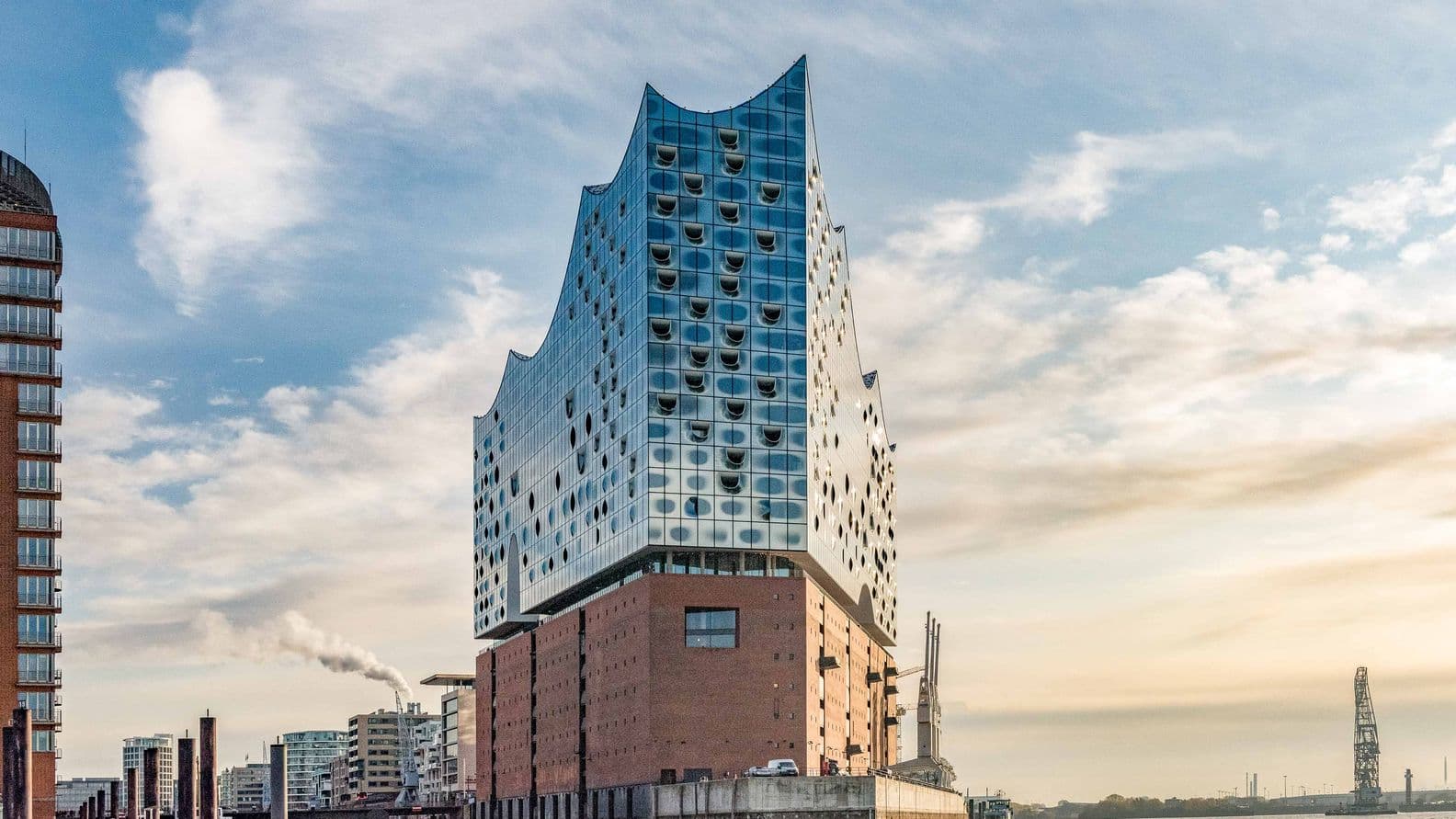 The Elbphilharmonie in Hamburg, a modern glass structure atop a brick base, stands by the water under a partly cloudy sky.