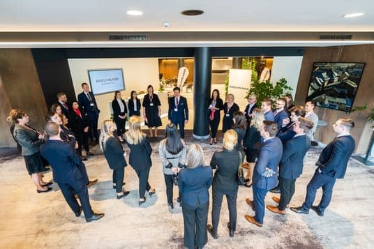 A group of people, including Engel & Völkers representatives and clients, standing in a circle during a social gathering in an elegant interior. Everyone is dressed formally, with company banners and logo in the background. The atmosphere feels both friendly and professional.