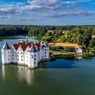 Luftaufnahme von Schloss Glücksburg mit roten Dächern, umgeben von einem See und üppigem Grün unter einem teilweise bewölkten Himmel.
