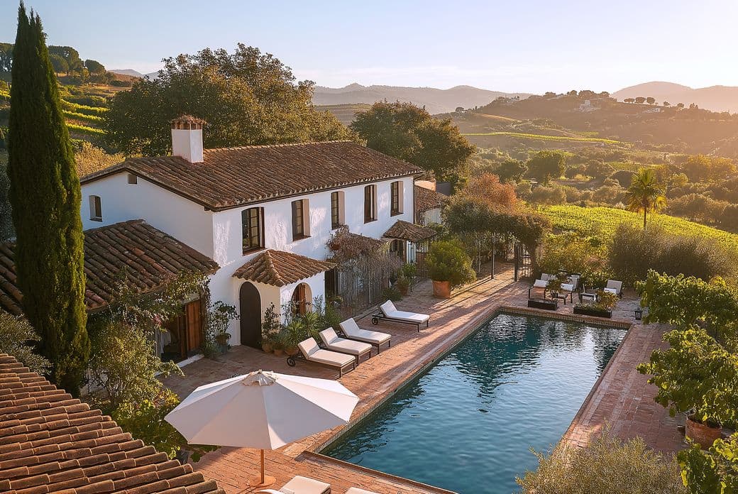 A picturesque villa with terracotta roof, surrounded by lush greenery, features a pool, lounge chairs, and an umbrella under a clear sky.