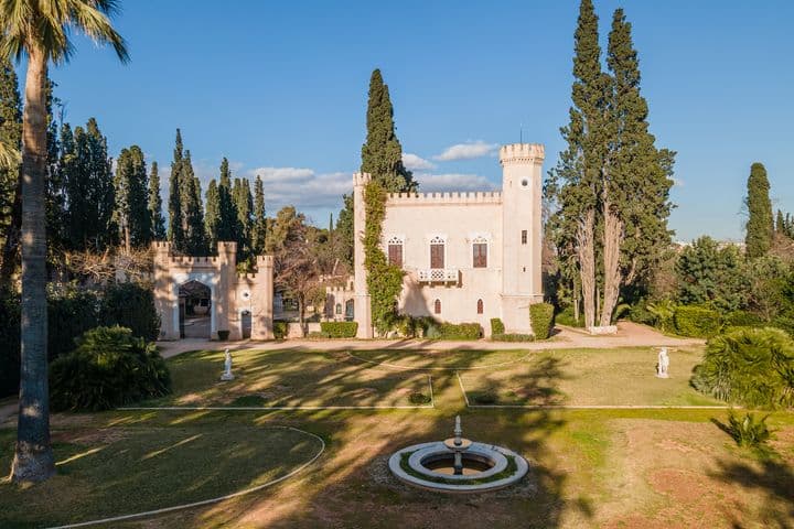 Vue aérienne d'un château rose clair avec une tour, entouré d'arbres verts et d'une fontaine au premier plan.