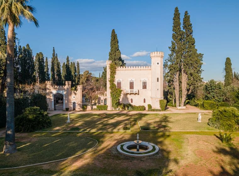 Aerial view of a light pink castle with a tower, surrounded by green trees and a fountain in the foreground.