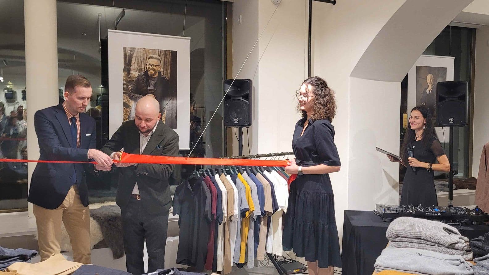 Two men cut a red ribbon inside a clothing store, with two women on each side — one holding the ribbon, the other speaking into a microphone.