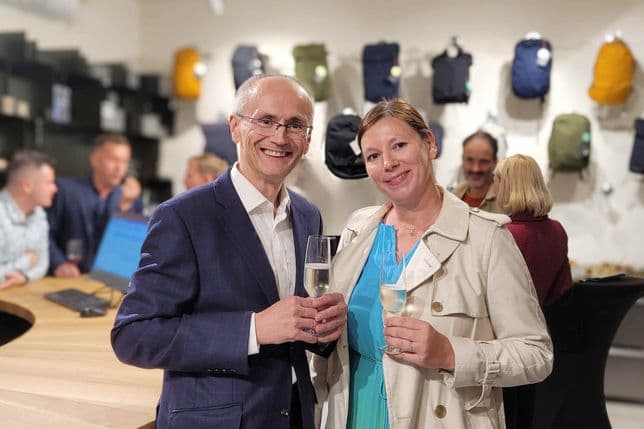 A man in a suit and a woman in a beige coat hold glasses of white wine and smile at the camera; in the background, a modern retail interior with backpacks and guests.