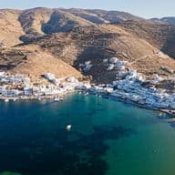 Aerial view of a coastal Greek village with white houses along a turquoise bay, small boats, and dry brown hills behind.