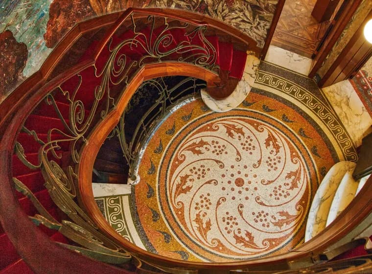 Ornate spiral staircase with red carpet and decorative wrought-iron railing, set on a colorful mosaic floor and marble walls.