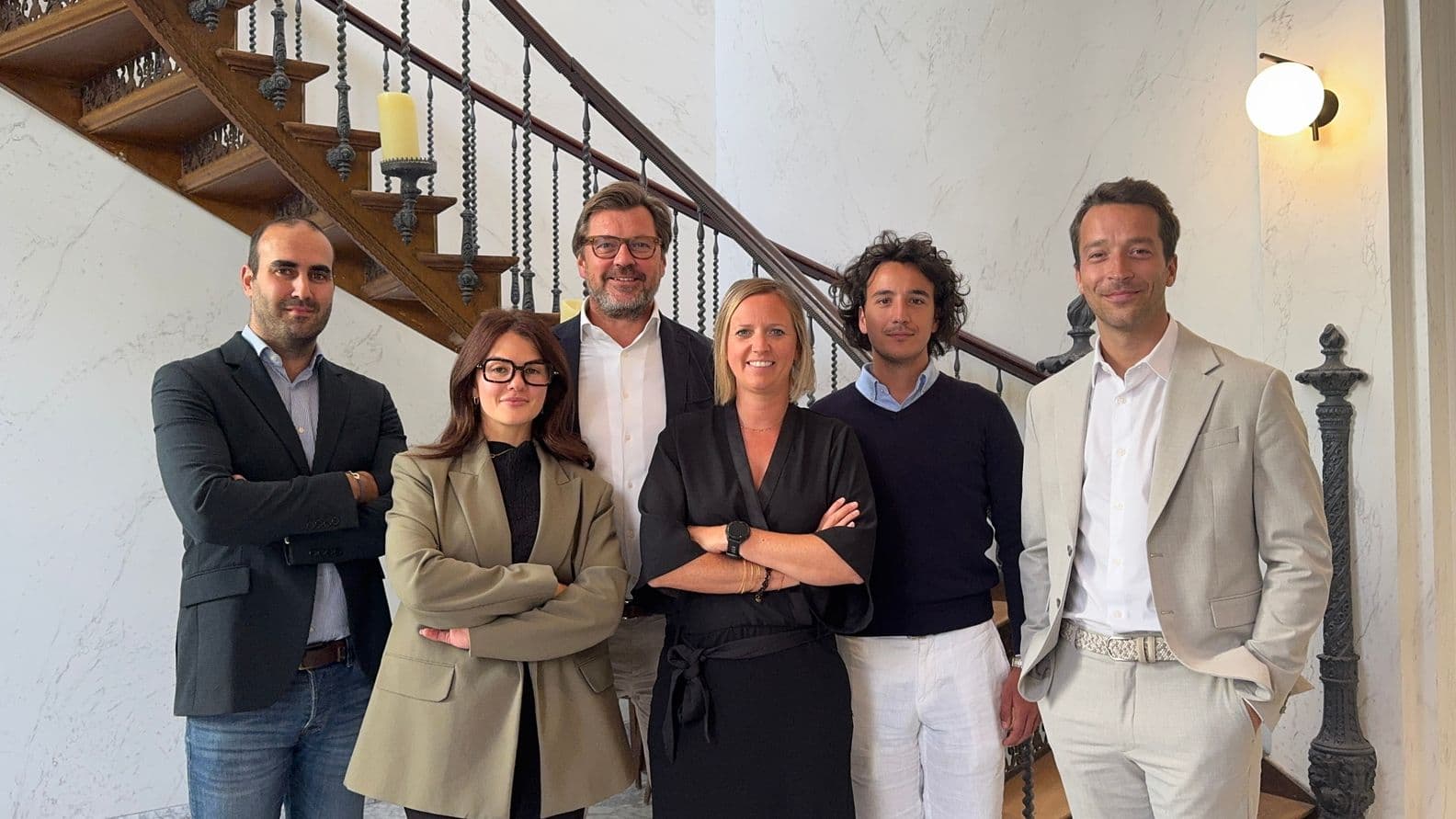 A group of six people standing in a hallway with a wooden staircase, dressed in business and casual attire, smiling at the camera.