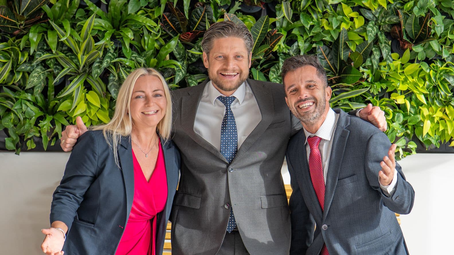 Three smiling people in suits standing close together with friendly gestures and arms around each other, set against a living green wall of plants.