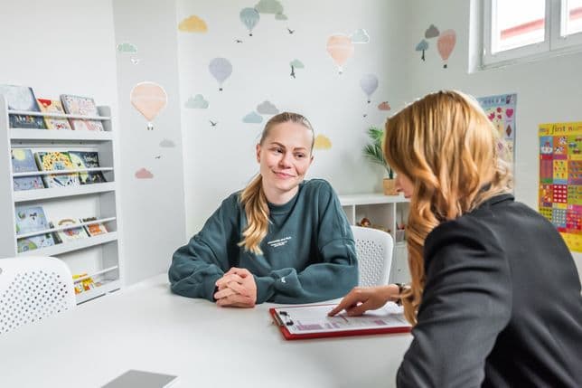 Two women sit at a table in a bright classroom. The school tenant on the left wears a sweatshirt and has a blonde ponytail; Eliška Tichá on the right, in a blazer, points to papers on a clipboard. Background includes balloon wall decals and a bookshelf with children’s books.
