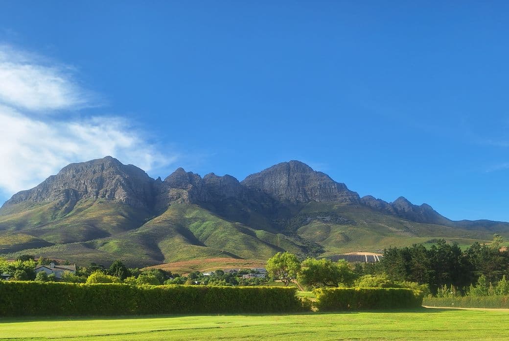 Scenic view of the Helderberg Mountains near Somerset West, South Africa. The dramatic mountain range with distinctive peaks rises above green foothills, with vineyards and residential properties visible in the middle ground. A manicured lawn is visible in the foreground, while the scene is captured under a bright blue sky with scattered clouds.