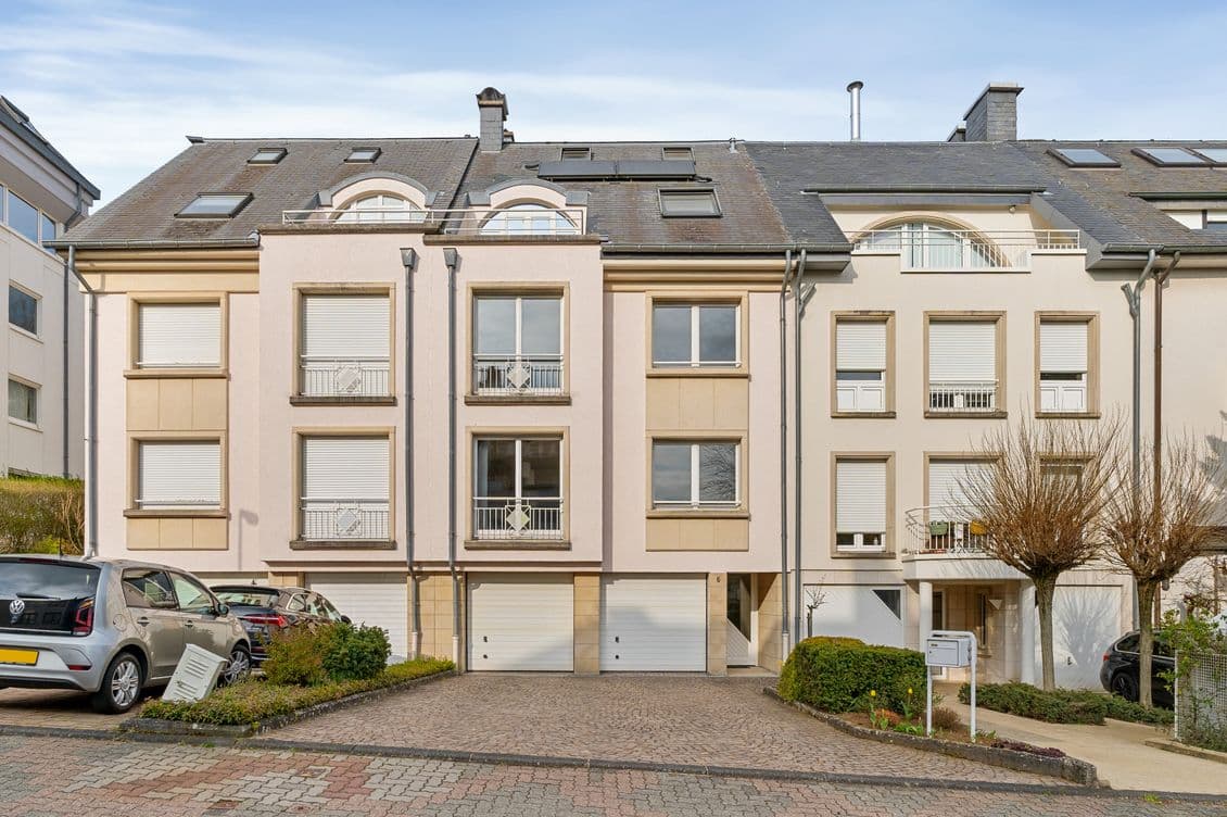 Row of beige, multi-story townhouses with slate roofs, arched dormer windows, balconies, and parked cars along a cobblestone street.
