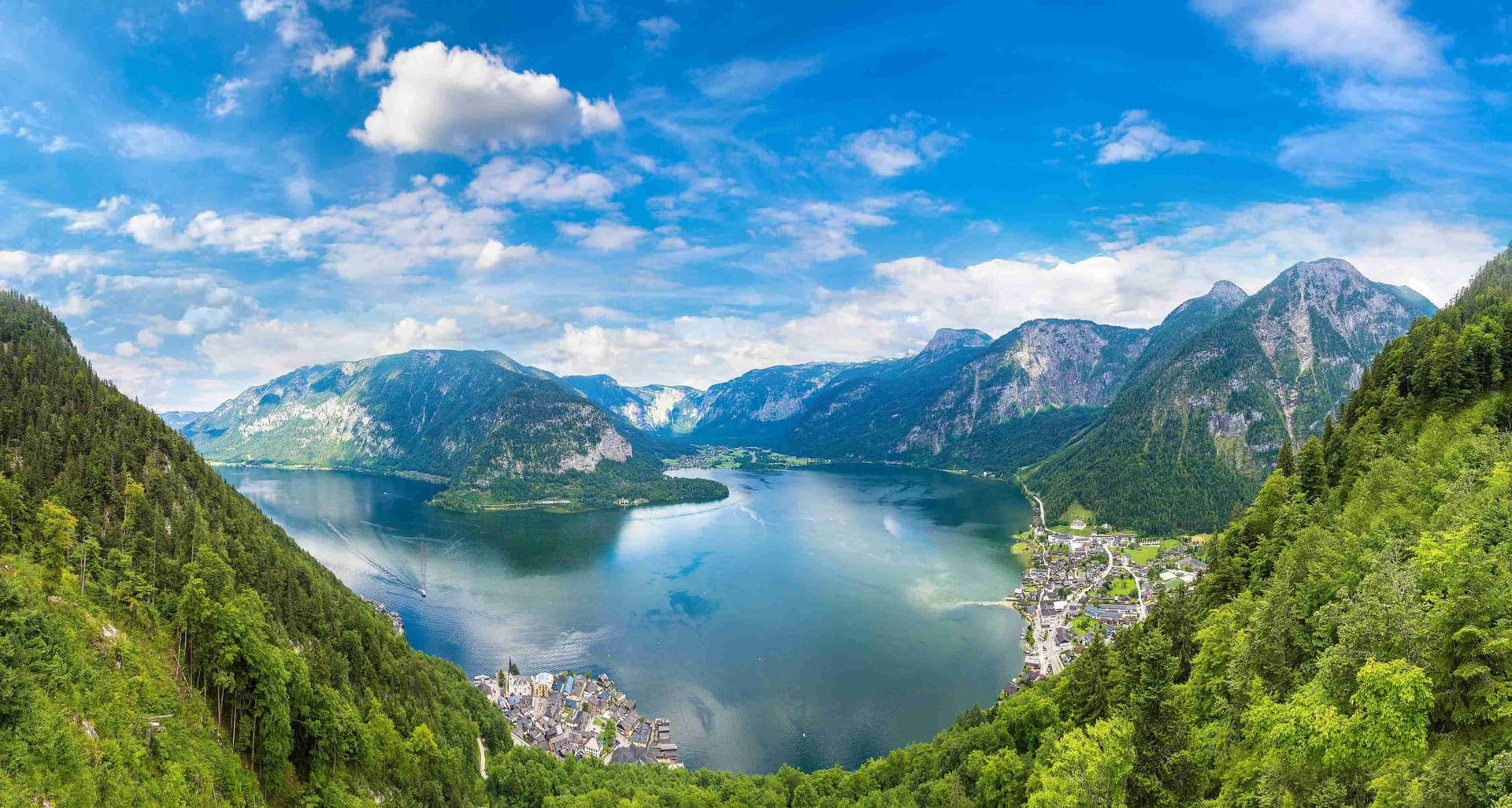 Panoramablick auf den gesamten Hallstätter See von oben, eingebettet zwischen steilen Bergwänden, mit Hallstatt und Obertraun an den Ufern unter blauem Himmel.