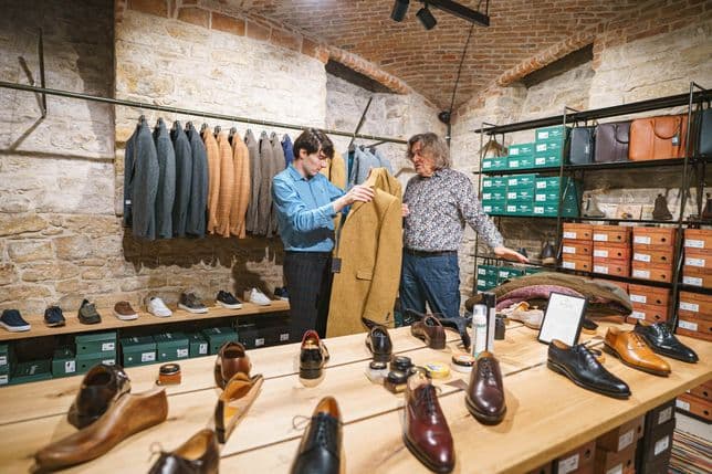 Two men examining a mustard-colored coat in a stylish men’s store with brick vaulted ceiling, stone wall, and shoes, suits, and leather accessories displayed on shelves and tables.