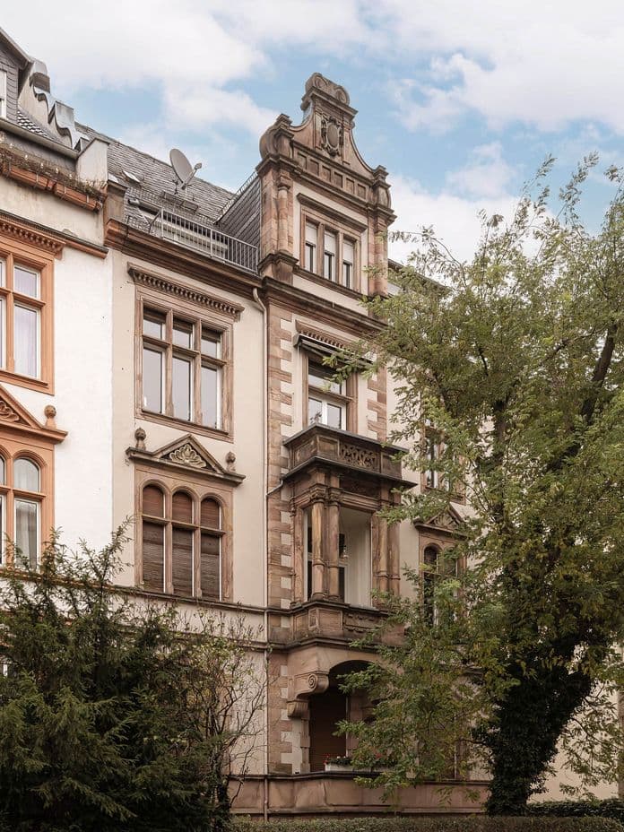 Ornate European residential buildings with decorative facades and large windows, adjacent to a leafy tree and greenery under a cloudy sky.