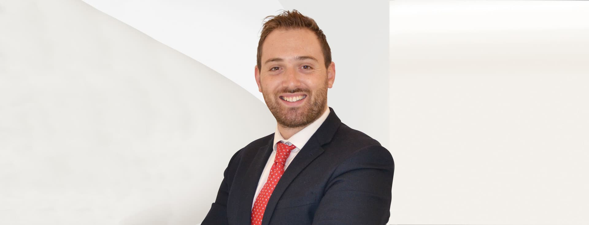 Smiling man in a suit with a red tie, standing against a plain white background.