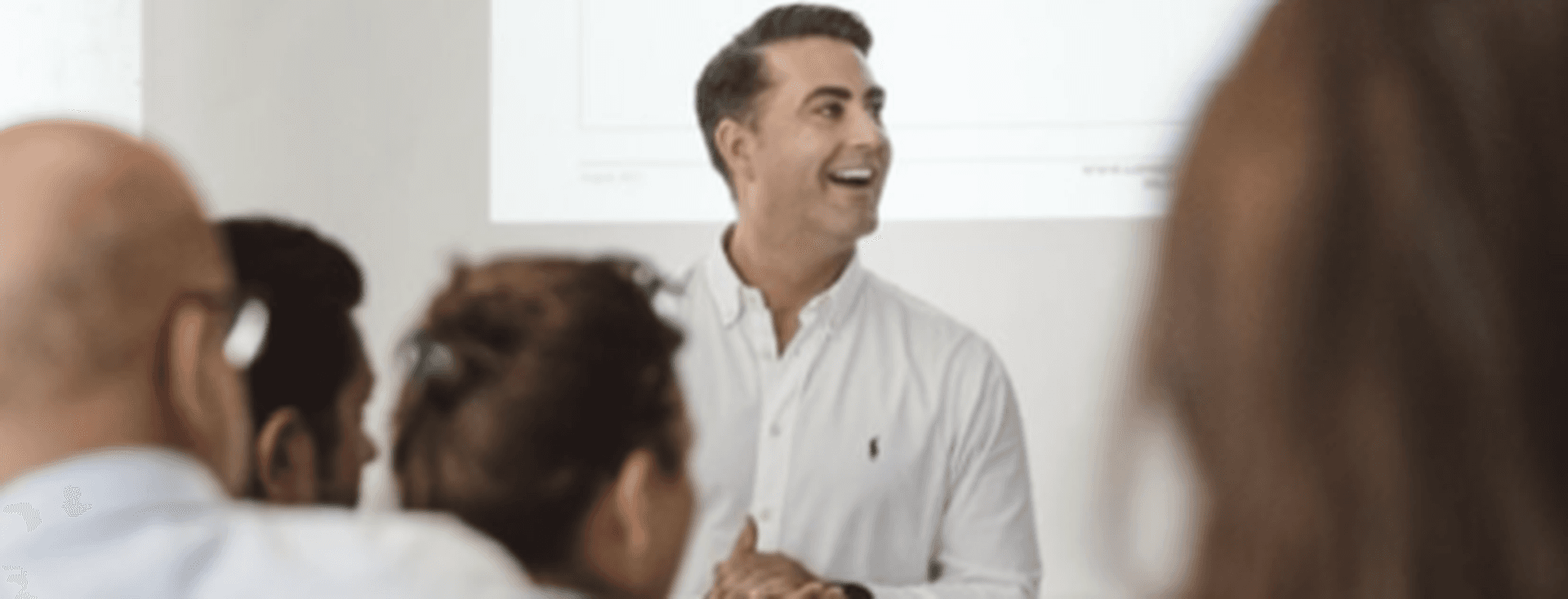 Un home somrient amb camisa blanca fa una presentació a un grup de persones davant d'una pantalla.