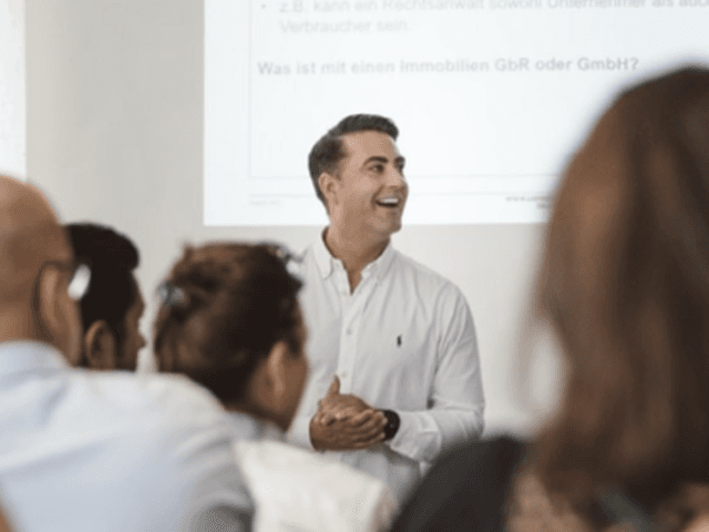 A person in a white shirt presenting to a group of colleagues in a bright office setting, gesturing enthusiastically while speaking to an attentive audience in a professional meeting environment.