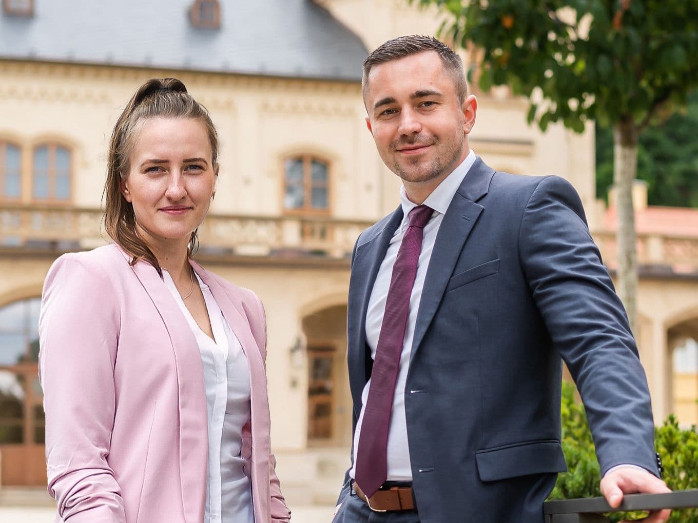 A man in a suit and a woman in a pink blazer stand outside in front of a historic building, both smiling at the camera.