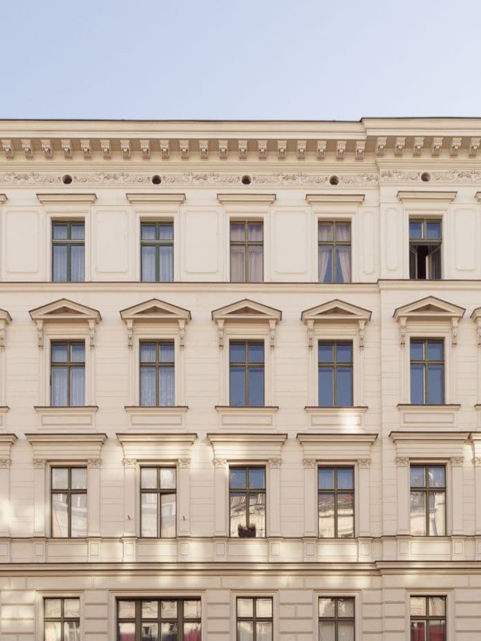Facade of a classic European building with ornate details, multiple windows, and a light beige color, flanked by similar structures.