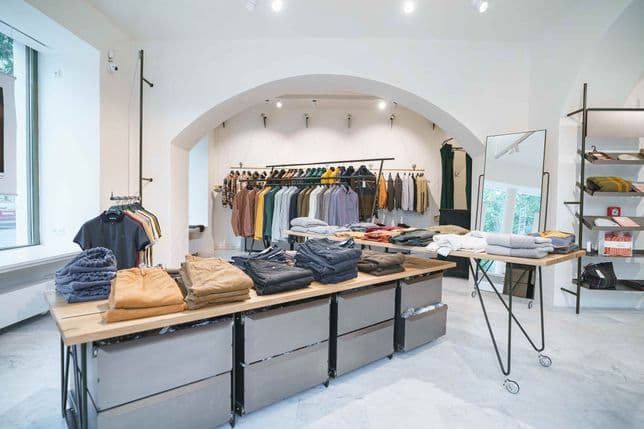 Interior of a modern men’s fashion store – folding tables with neatly stacked trousers and sweaters, background clothing racks with shirts and jackets, bright airy space with arched ceiling.