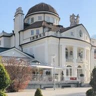 Elegante castello storico con cupola, circondato da alberi curati e montagne sullo sfondo, sotto un cielo azzurro e limpido.