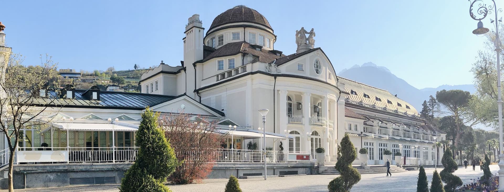 Elegantes historisches Schloss mit Kuppel, umgeben von gepflegten Bäumen und Bergen im Hintergrund, unter einem klaren blauen Himmel.