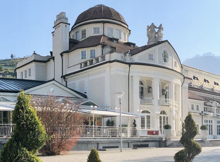 Elegantes historisches Schloss mit Kuppel, umgeben von gepflegten Bäumen und Bergen im Hintergrund, unter einem klaren blauen Himmel.