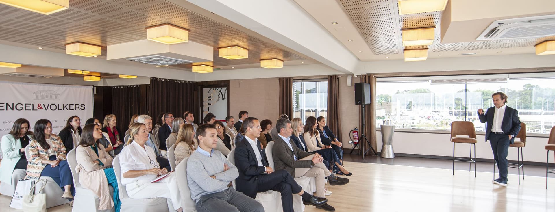 Un agent immobiliari presenta davant d'un públic assegut en una sala lluminosa amb bosses de mà i el logotip d'Engel & Völkers.