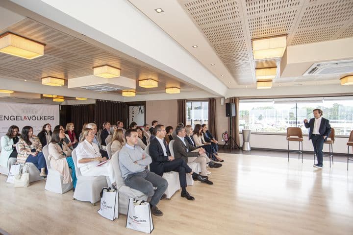 Un agente inmobiliario presenta a un público sentado en una sala luminosa con bolsas de mano y el logotipo de Engel & Völkers.