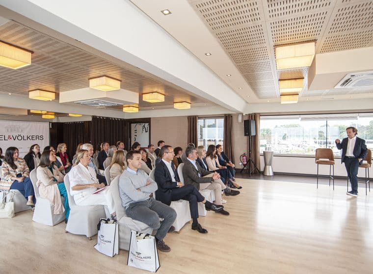 Un agent immobiliari presenta davant d'un públic assegut en una sala lluminosa amb bosses de mà i el logotip d'Engel & Völkers.