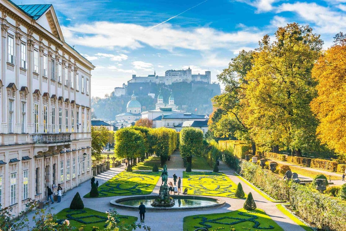 Mirabellgarten im Herbst mit barockem Schloss Mirabell, geometrischen Blumenbeeten, Springbrunnen und Festung Hohensalzburg im Hintergrund unter blauem Himmel.