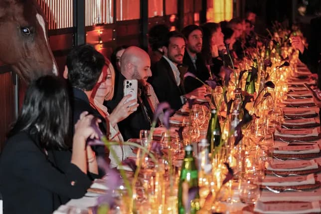 Long banquet table with guests in formal attire dining by candlelight; a horse peeks from a stable on the left.