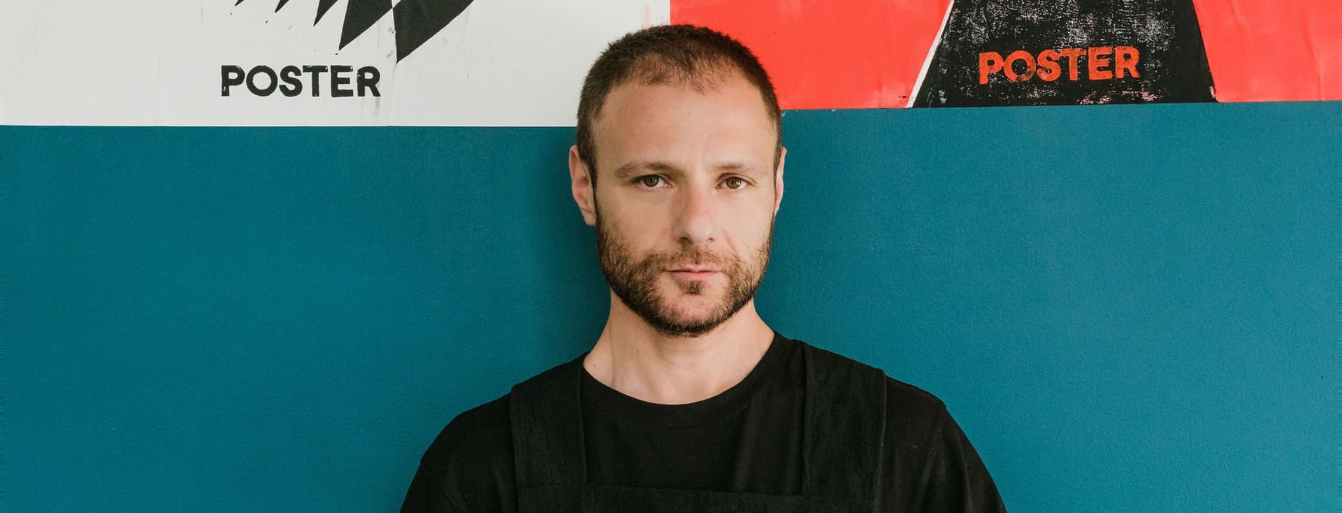 Ένας άντρας με γένια στέκεται μπροστά σε έναν μπλε τοίχο με αφίσες.