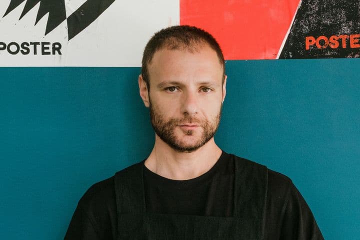 Ένας άντρας με γένια στέκεται μπροστά σε έναν μπλε τοίχο με αφίσες.