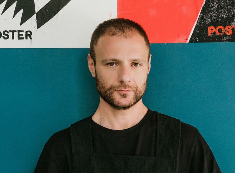 Ένας άντρας με γένια στέκεται μπροστά σε έναν μπλε τοίχο με αφίσες.