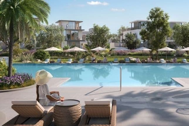 Woman in a sun hat sitting on a lounge chair by a pool, surrounded by greenery and modern buildings, under a clear blue sky.