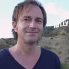Smiling man with light brown hair wearing a navy V-neck shirt, outdoors with rocky hills and a partial Hollywood sign in the background.