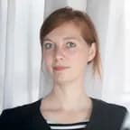 Portrait of a young woman with light brown hair and blue eyes, wearing a black top, standing before white curtains.