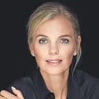 Nahaufnahme einer blonden Frau mit blaugrünen Augen, die ein dunkles Shirt trägt und vor einem dunklen Hintergrund sanft lächelt.
