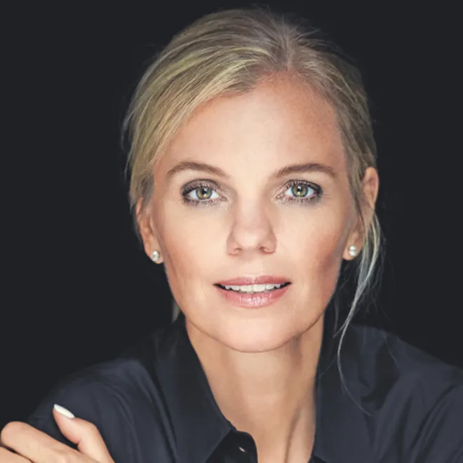 Close-up portrait of a blonde woman with blue-green eyes, wearing a dark shirt, smiling softly against a dark background.